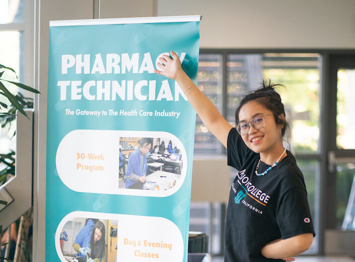 Pharmacy Technician standing banner. Young Asian-American woman in black tshirt and glasses stands next to it and gestures.