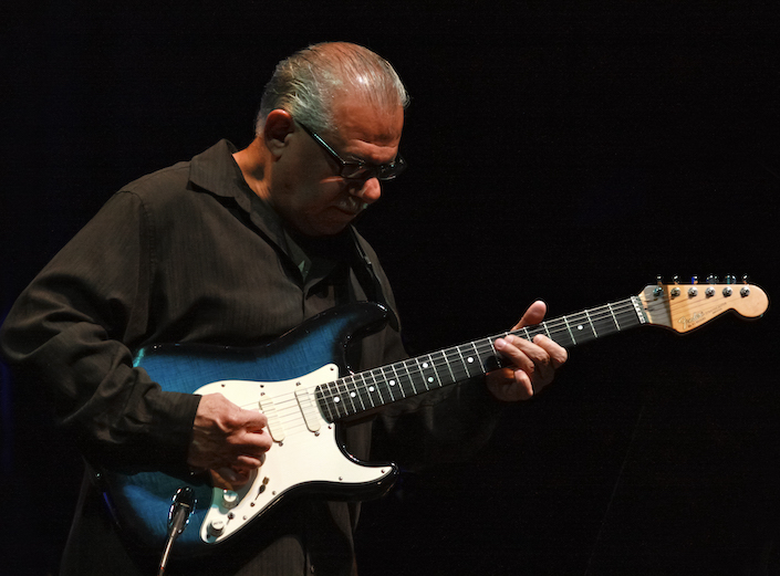 Ray Obiedo, a Latin Jazz guitarist performing in dark clothes against a black background. He is an older man, with short grey hair and glasses.