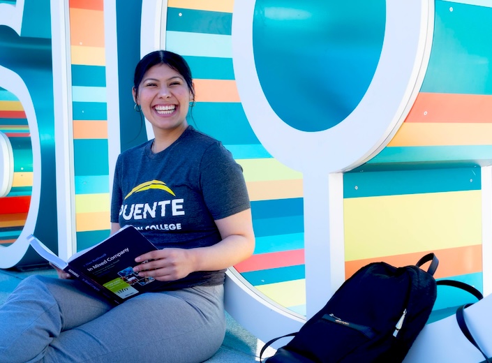 Puente student, a twenty-something Latina in a navy blue Puente tshirt smiles and reads a textbook.